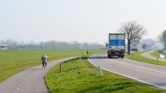 A cycle path in the Dutch countryside. A road with large lorries on it, with a bi-directional cycleway on both sides of the road, each separated by a large grassed area.