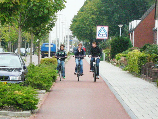 Three boys riding side by side on a red asphalt cycleway, separated from motor traffic by bushes, and with a forgiving, angled kerb up to the footway.