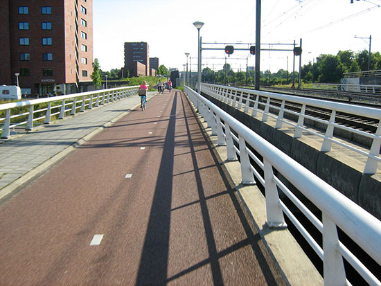 A bridge across a major road. To the left is a footway, and we can see the wide bi-directional cycleway. There are street lights too. To the right we can see a parallel railway bridge.