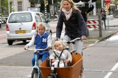 A mother rides a bakfiets (box-bike) in which her youngest daughter sits. Her older daughter rides her own bike alongside.