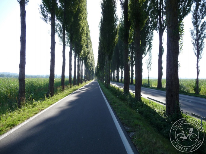 A wide, flat cycle path alongside a rural road, but separated from the road by grass and trees.