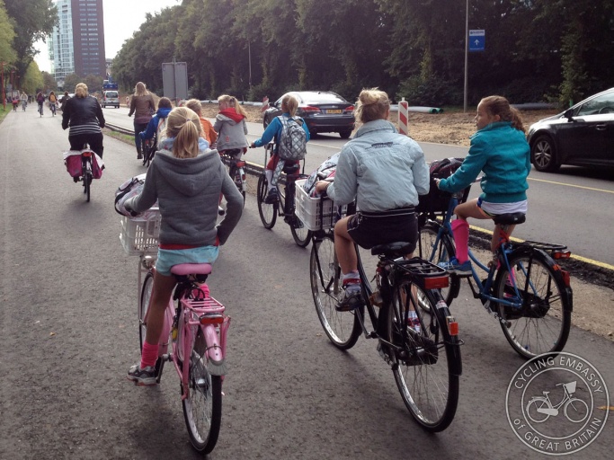 A wide cycle path alongside (but physically separate from) a road. A group of children ride bikes, their bags in the crates attached to the front.