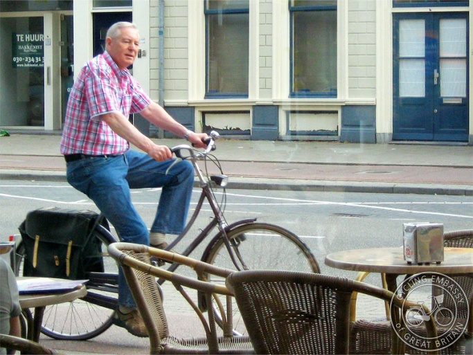 A middle-aged man rides his bike along a cycle path past a café
