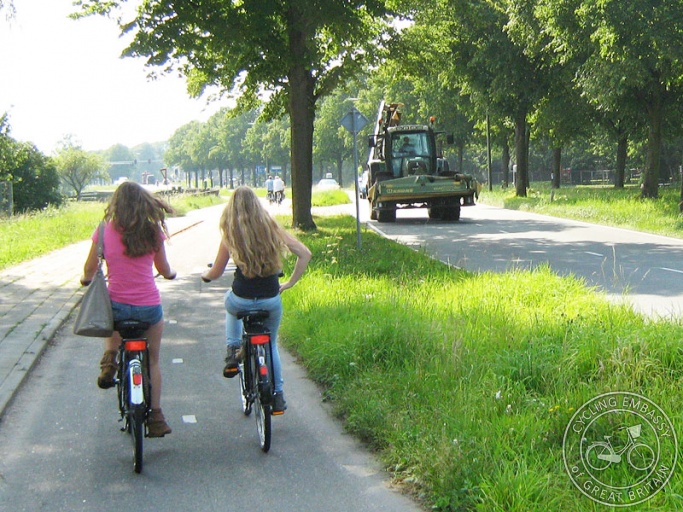 Two girls use a cycle path which is separated from the busy road by a wide grass verge and trees